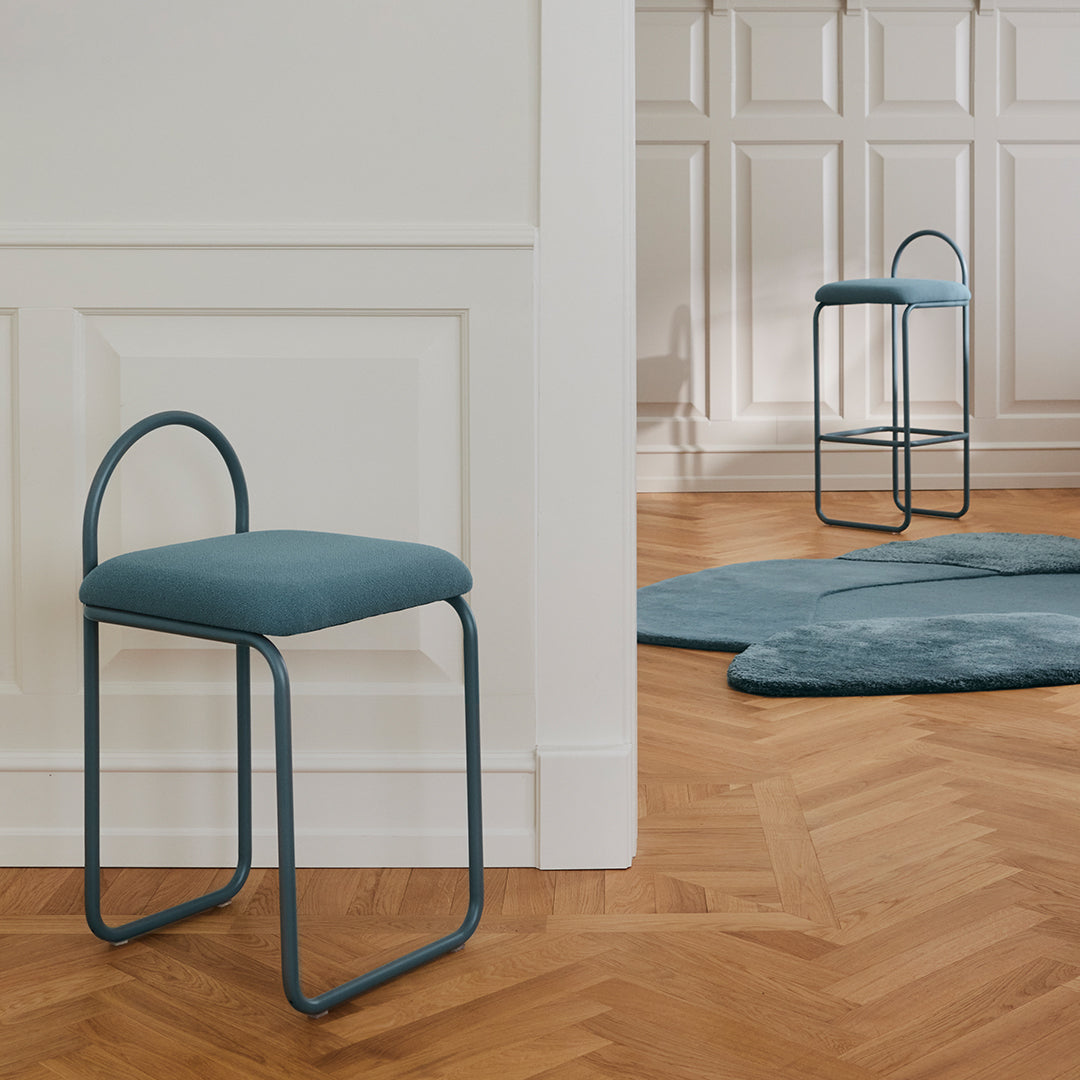 Teal upholstered stools with curved metal frames against a warm herringbone wooden floor and a soft, blue rug.