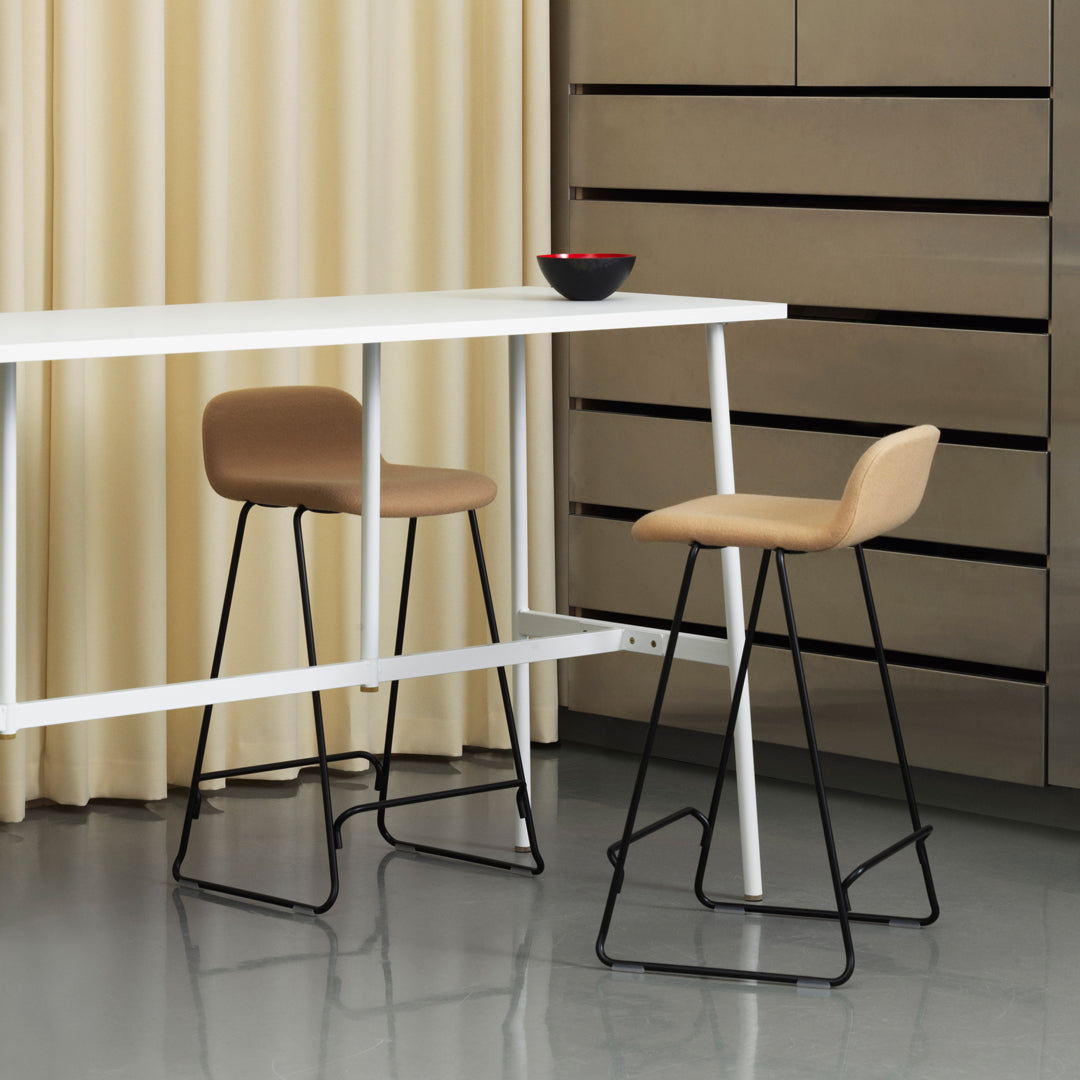 "Neutral beige stool with black legs at a modern white bar table, against soft cream drapes and sleek grey flooring."