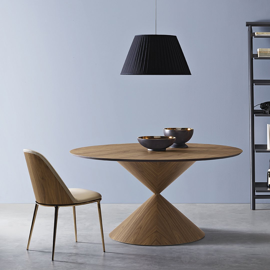 Round wooden dining table in natural tones, paired with a beige chair and a black pleated pendant light against a blue wall.