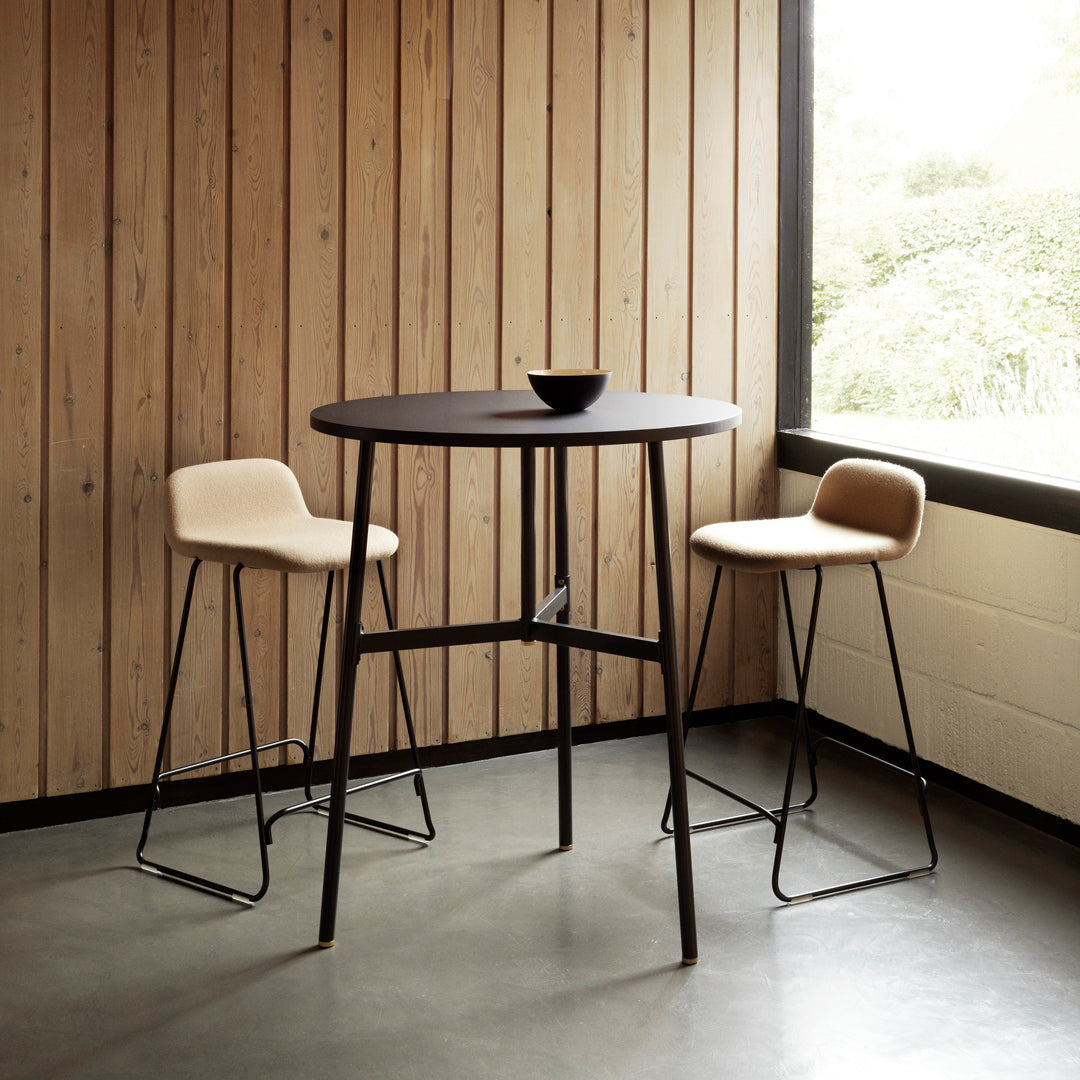 Modern black round table with minimalist design, paired with cream upholstered bar stools against wood-paneled wall.