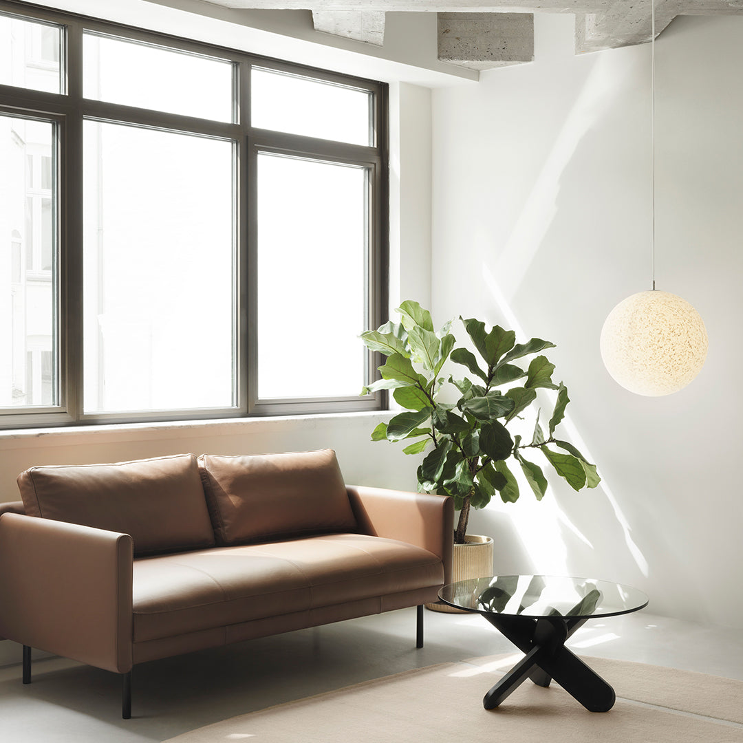 Brown sofa with smooth texture, glass coffee table, green plant, and warm pendant light in a bright, minimalist space.