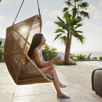 "Light brown wicker hanging chair with gray cushions, surrounded by green palm plants and a serene outdoor setting."