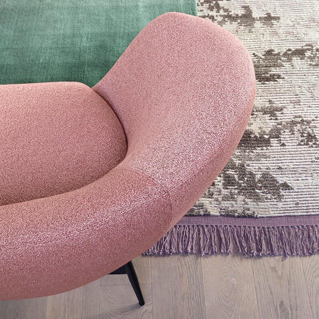 Curved pink chair with textured fabric, set against a green rug and patterned fringe. Modern and stylish design.
