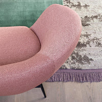Curved pink chair with textured fabric, set against a green rug and patterned fringe. Modern and stylish design.