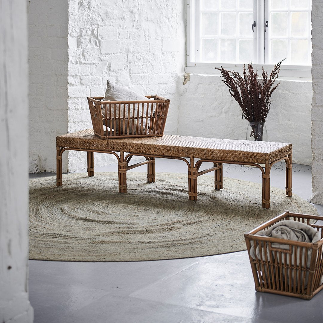 "Natural rattan bench with a beige cushion, set on a round, textured green rug in a minimalist interior."