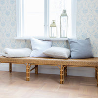 Neutral-toned pillows in white, beige, and light gray on a woven bench against a patterned blue wallpaper backdrop.