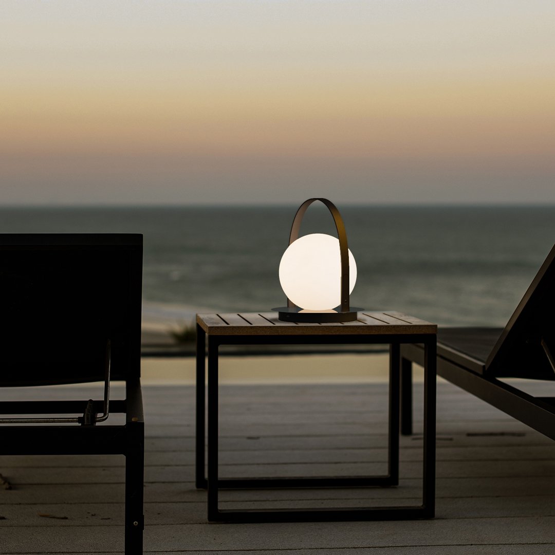 Modern white orb lamp with sleek black handle, set against a tranquil sunset backdrop and ocean hues.