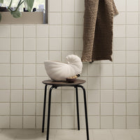 White spiral decorative pillow on a black stool, with a textured brown throw against a minimalist tiled wall.
