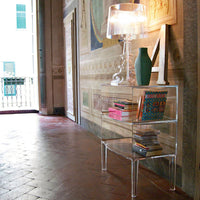 Clear acrylic console table with stacked colorful books, teal vase, and a stylish lamp in a vibrant hallway setting.