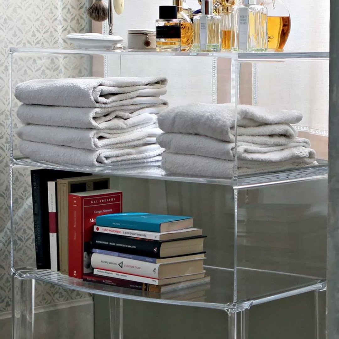 Stacked grey towels with a smooth texture on a clear acrylic shelf, alongside colorful books and elegant glass bottles.