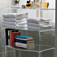 Stacked grey towels with a smooth texture on a clear acrylic shelf, alongside colorful books and elegant glass bottles.