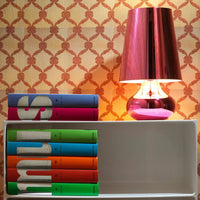 Colorful stacked books with bold patterns against a geometric wallpaper and a shiny pink lamp.