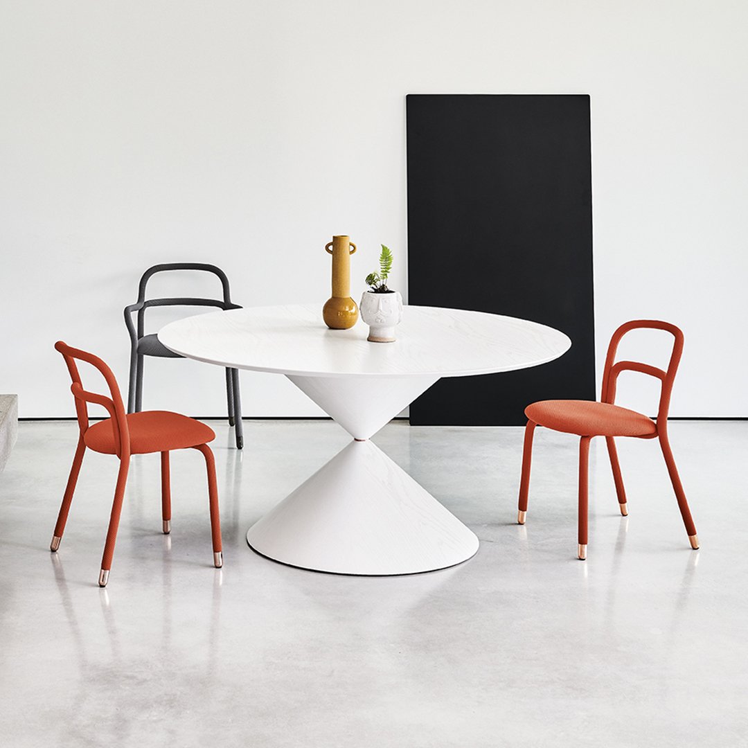 Modern round white dining table with a sleek cone base, paired with orange and black chairs against a minimalist backdrop.