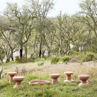 Natural terracotta stools in various sizes, featuring smooth curves and earthy tones, set against a lush green backdrop.