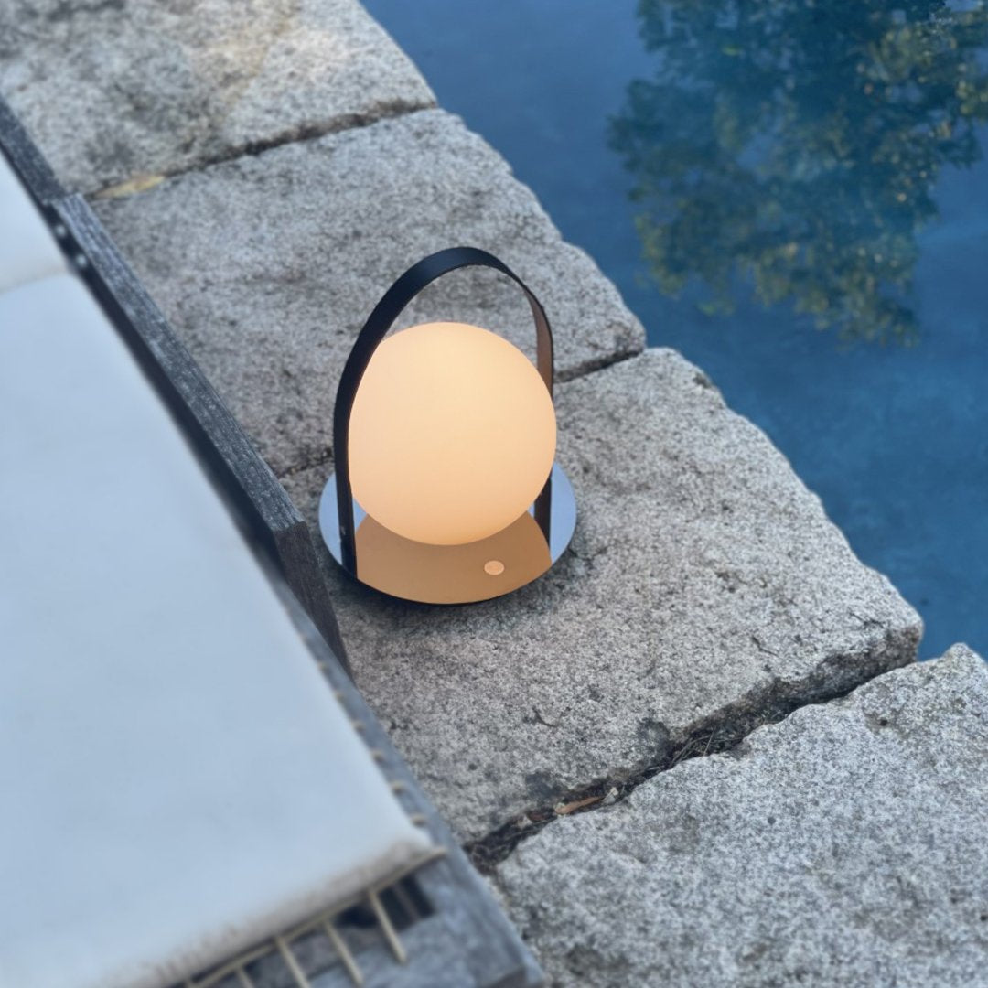 Round white lamp with a sleek black handle, set on textured gray stone by a tranquil blue pool.
