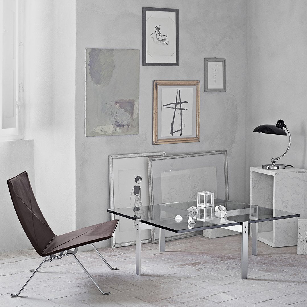 Elegant glass coffee table with sleek chrome legs, paired with a brown lounge chair against a minimalist gray backdrop.