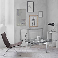 Elegant glass coffee table with sleek chrome legs, paired with a brown lounge chair against a minimalist gray backdrop.