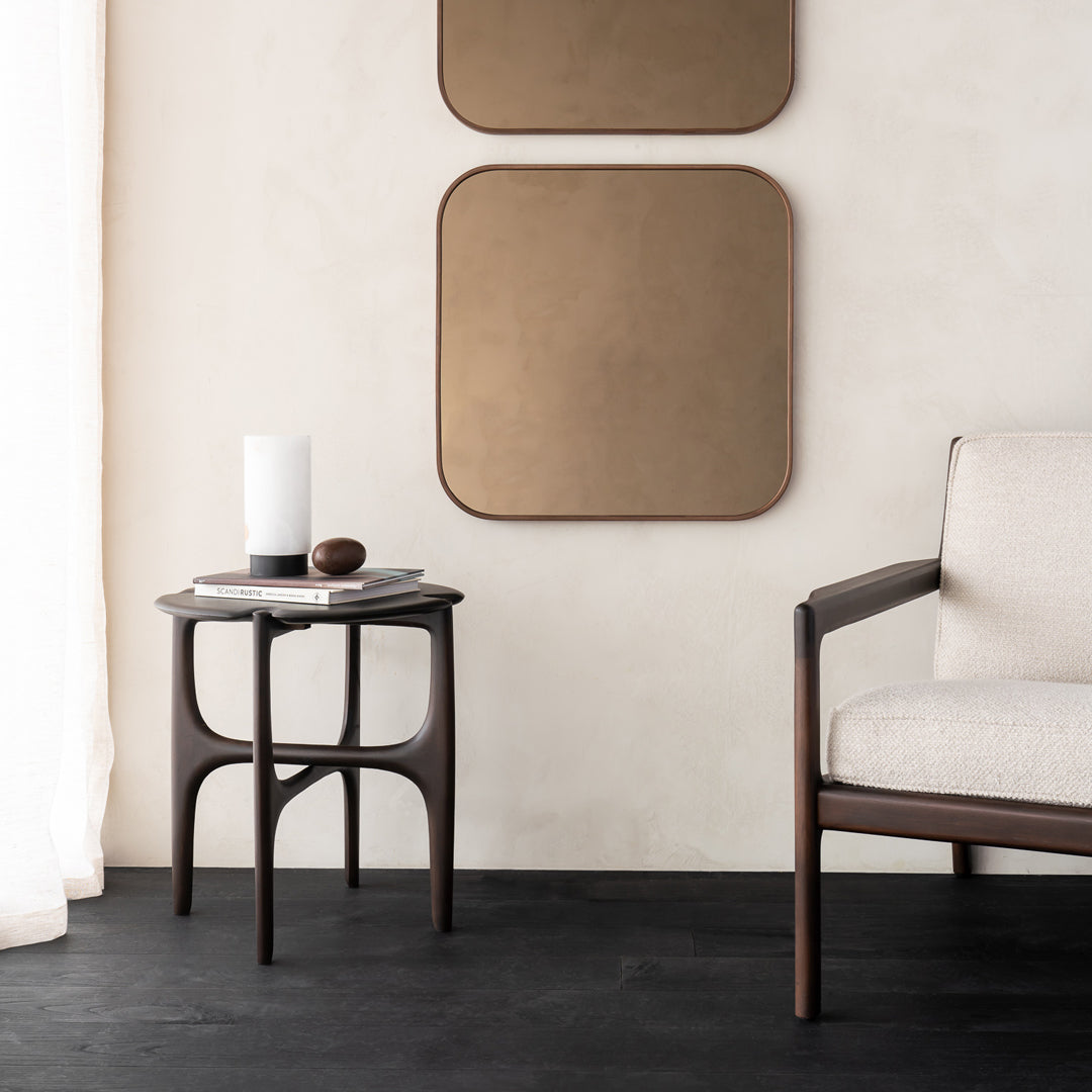 Walnut side table with smooth lines, paired with a beige chair, set against a light textured wall and dark floor.