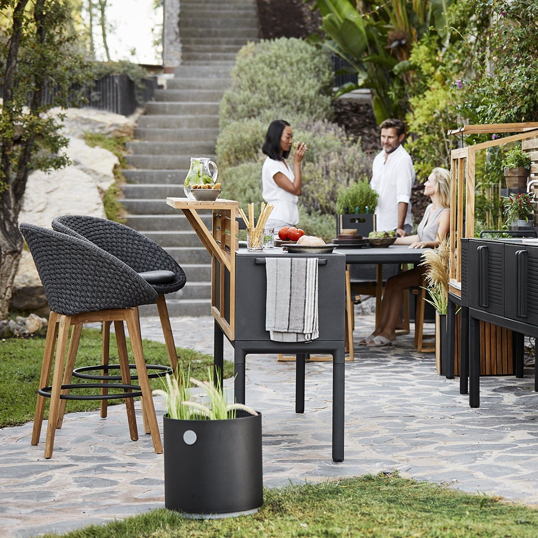 Black woven bar stools, sleek black table, wooden accents, and green plants set in a natural outdoor stone patio.
