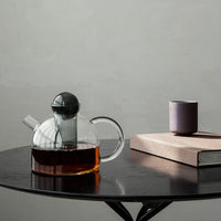 Clear glass teapot with black lid, amber tea, and a minimalist purple cup on a sleek black table.
