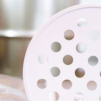 Light pink round drainer with circular holes, set against a soft, blurred metallic background.