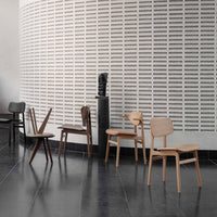 Assorted wooden chairs in natural, dark brown, and black finishes against a textured white wall. Modern minimalist design.