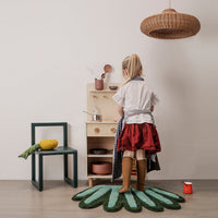 Child playing in red skirt and brown boots on a green leaf-patterned rug, with minimalist wooden kitchen setup.