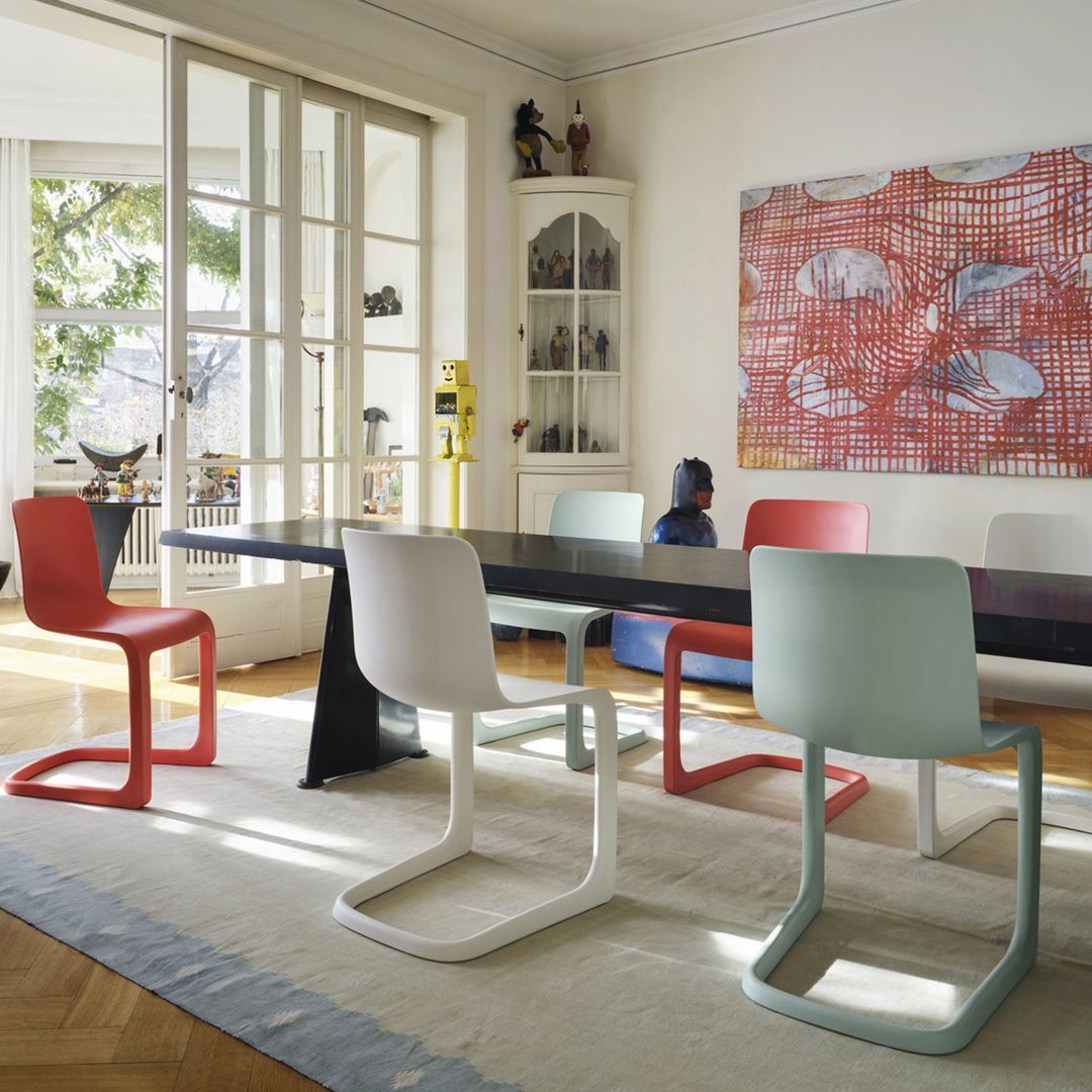 Colorful modern chairs in red, mint, white, and pink arranged around a sleek black dining table, set in a bright room.