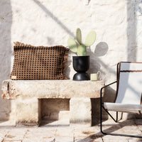 Brown polka dot decorative pillow with fringed edges on a light stone bench, paired with a black planter and beige chair.