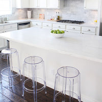 "Modern kitchen scene with white marble countertop, clear acrylic stools, and green apples in a bowl."