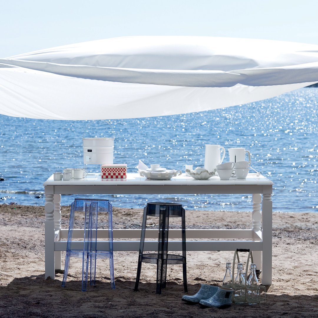 White table with transparent blue and black stools, set against a serene ocean backdrop and bright white fabric.