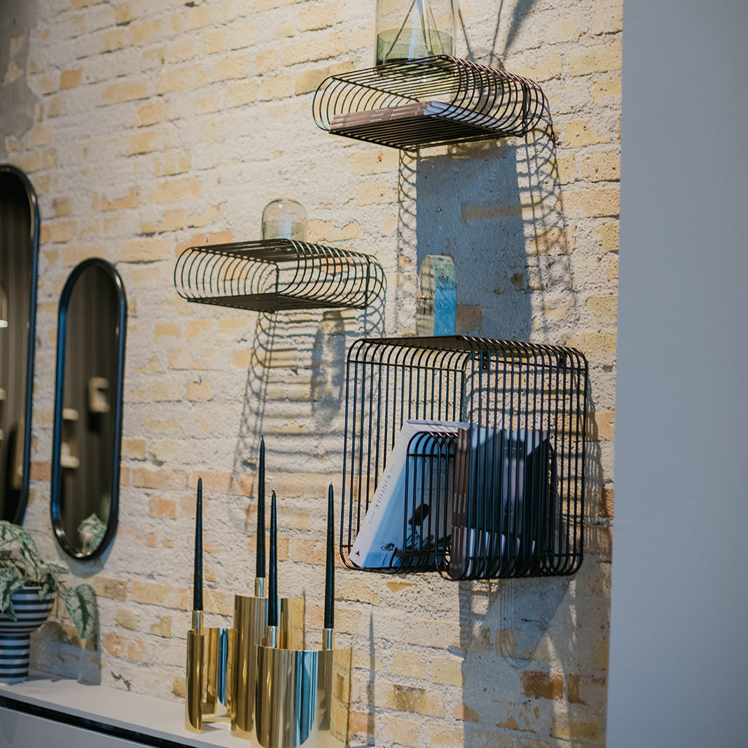 Black wire wall shelves with green glass accents and decorative gold candle holders against a textured beige brick wall.