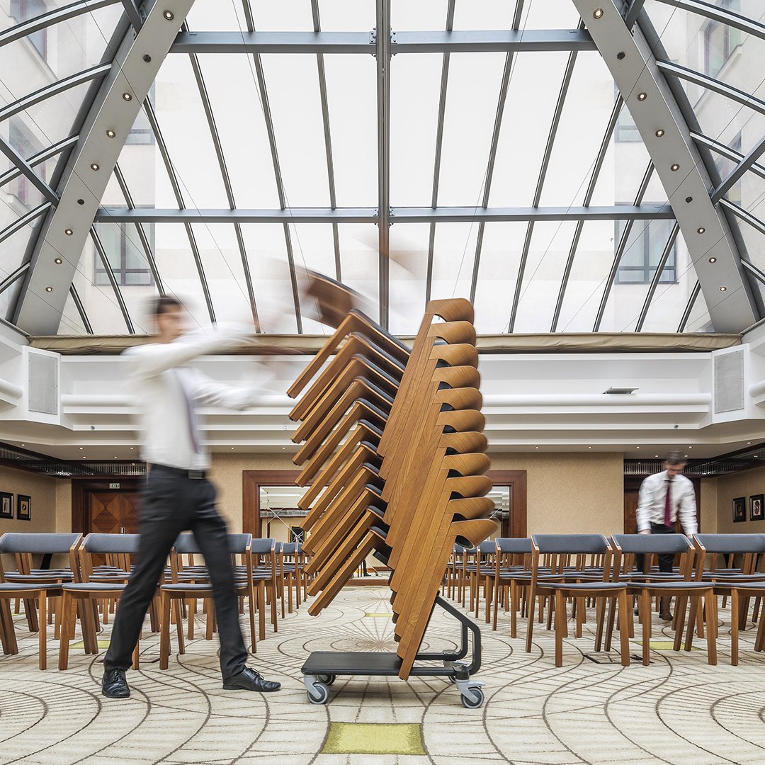 Stacked wooden chairs with a warm brown finish, set against a modern, light-toned patterned floor.