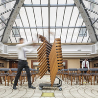 Stacked wooden chairs with a warm brown finish, set against a modern, light-toned patterned floor.