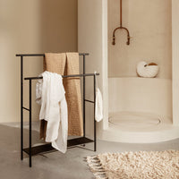 Black towel rack with beige and white towels, set against a warm neutral background and subtle textured rug.
