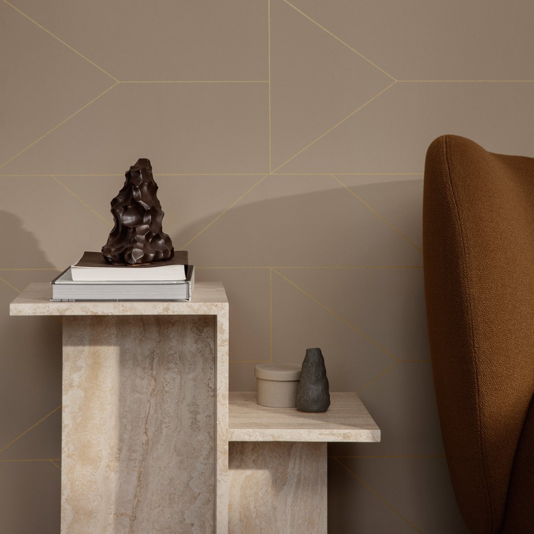 Minimalist beige wall with geometric patterns, featuring a light stone table and warm brown armchair.