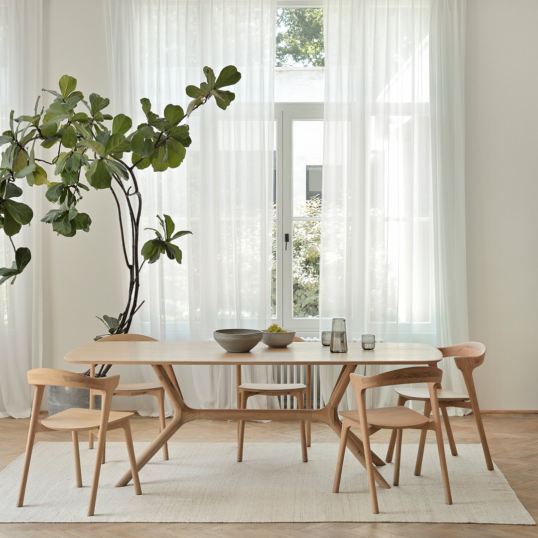Light wooden dining table with natural grain, surrounded by beige chairs in a bright, airy room with sheer curtains.