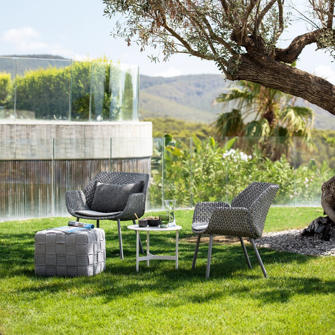 Gray woven patio chairs and a cube ottoman on green grass, surrounded by natural greenery and a modern backdrop.