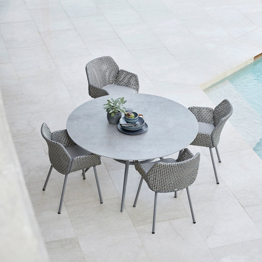 Gray round table with textured gray wicker chairs, set on a light stone patio near a pool.