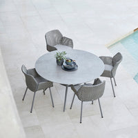 Gray round table with textured gray wicker chairs, set on a light stone patio near a pool.