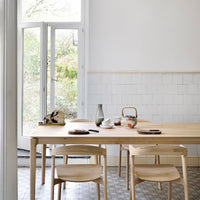 Light wood dining table with natural tones, paired with simple beige chairs and a white wall, featuring subtle grey tile patterns.