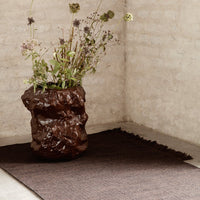 Textured brown vase with dried flowers on a dark brown woven rug against a light beige wall.
