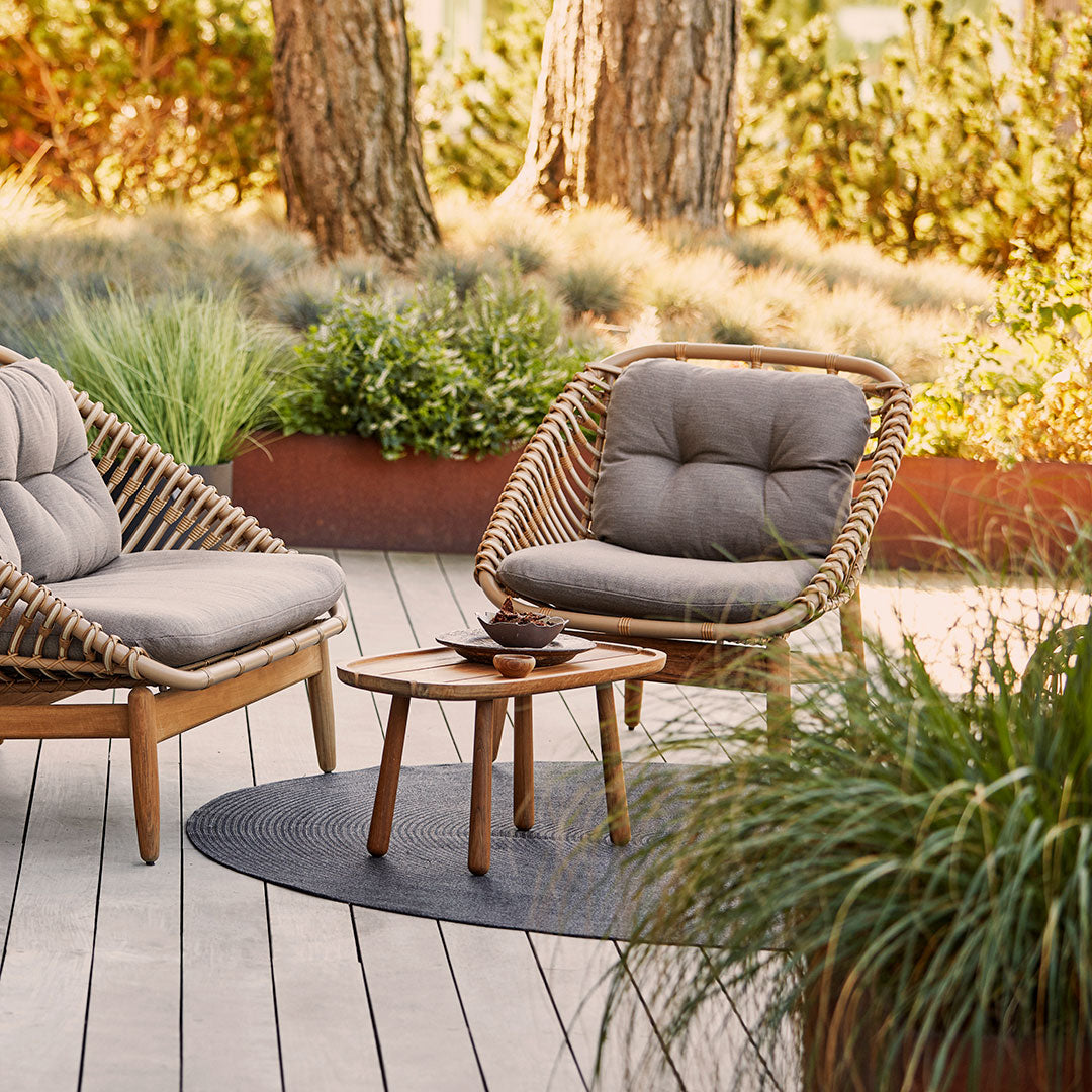 Light gray chairs with natural wood frames, paired with a round dark woven rug, set against lush greenery.