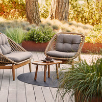 Light gray chairs with natural wood frames, paired with a round dark woven rug, set against lush greenery.