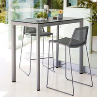 Gray square table with sleek metal legs and matching woven bar stools in a contemporary indoor setting.