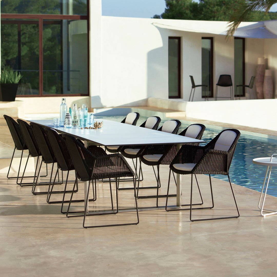 Black woven chairs around a sleek white dining table by a pool, with a neutral stone patio backdrop.