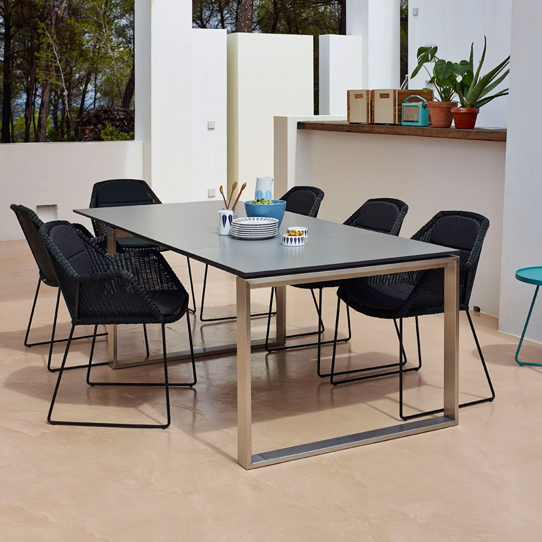 Outdoor dining table with sleek gray surface and black woven chairs, set against a neutral earth-tone background.