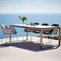 Stainless steel dining table with light gray wicker chairs, set against a vibrant blue sea backdrop. Outdoor elegance.