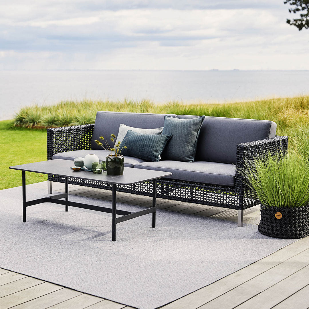 Black wicker sofa with gray cushions and a matching coffee table on a light gray outdoor rug. Green plants in a black pot.