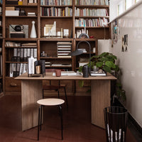 Modern wooden desk with light oak finish, black accents, and green plant against a backdrop of bookshelves and neutral walls.
